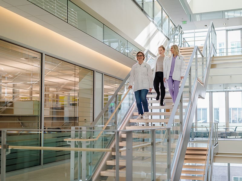 Three professionals in lab coats walk down a modern glass staircase in a bright, open research facility, engaged in conversation.