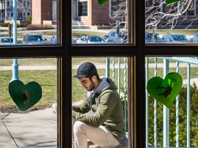 A student wearing a cap and earbuds studies outside, viewed through a window decorated with green heart-shaped notes featuring Spartan helmet drawings.