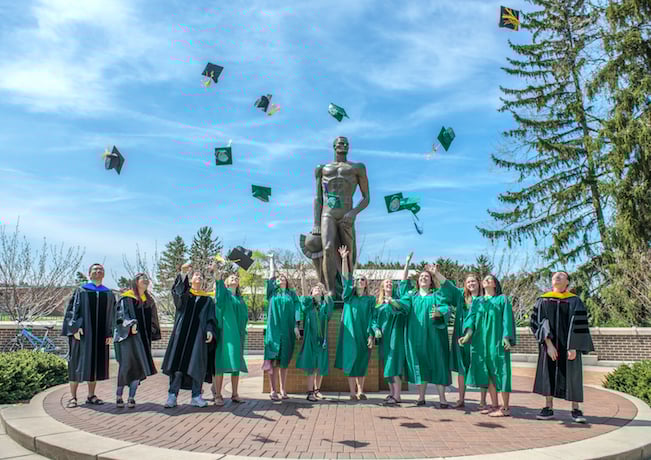 Undergraduates in green MSU robes toss their caps into the air around Sparty statue.