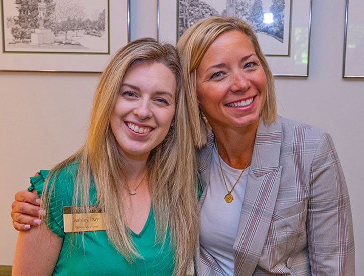 Ashley Day, event coordinator, left, and Heather Fink, CMP and senior university academic events planner, right, smile as they pose for a picture. Ashley is wearing a green shirt with a gold name badge, and Heather is wearing a gray plaid blazer with a gold necklace.