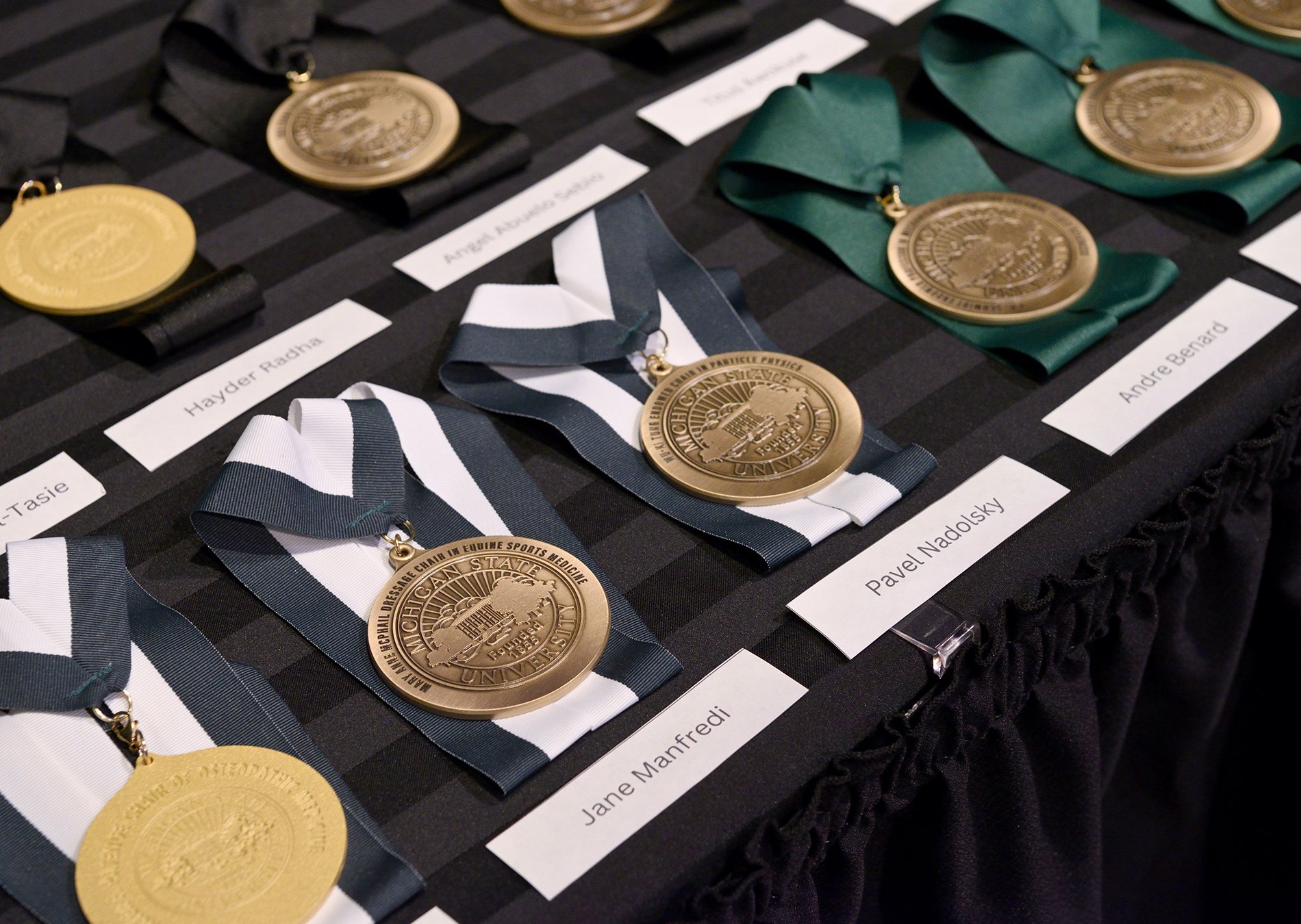 Two rows of gold-colored medals with green, black and white, and black ribbons sit against a black tablecloth.