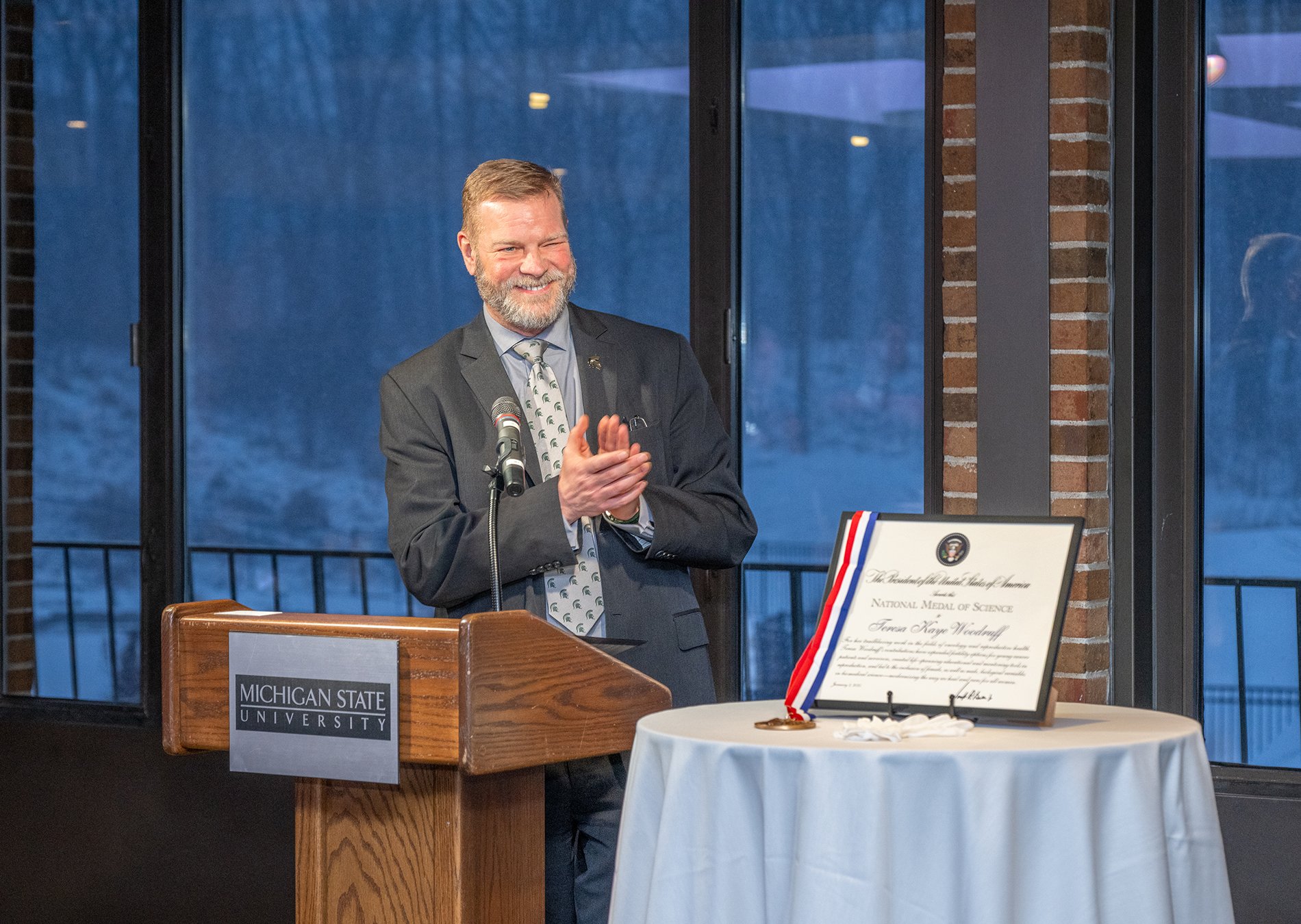 Interim Provost Jeitschko applauds behind a podium, Teresa Woodruff's National Medal of Science award and medal displayed on a table beside him.