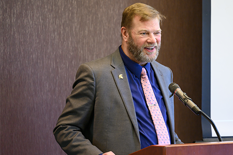 Thomas Jeitshcko standing at a podum speaking into a microphone. He is wearing a grey suit jacket, a red tie and a blue shirt with a spartan helmet pin on his right lapel.