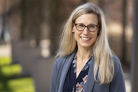 Portrait of Laura Lee McIntyre, photographed outside, wearing a blue-grey suit jacket over a dark floral blouse. Her blonde hair is down over her left shoulder and she is wearing glasses while smiling at the camera. 