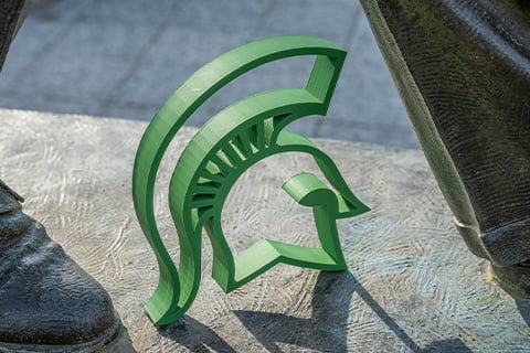 A green 3D printed spartan helmet sits in between the metal feet of the Hannah statue on MSU's campus.