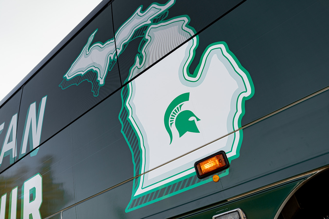 Side of passenger bus showing the outline of the state of Michigan with a green Spartan helmet in the middle of the lower peninsula. 
