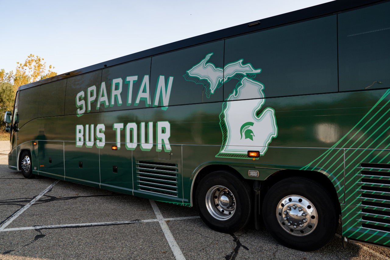 Side of a passenger bus with green skin, says Spartan Bus Tour and an outline of the state of Michigan with a Spartan helmet in the middle of the lower peninsula.