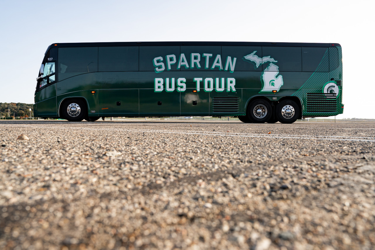 Side of a passenger bus with green skin, says Spartan Bus Tour and an outline of the state of Michigan with a Spartan helmet in the middle of the lower peninsula, seen from across a paved lot. 