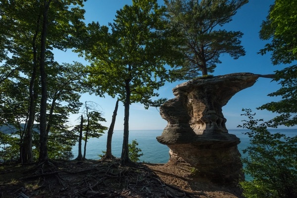 A rock formation surrounded by trees with a lake and blue sky in the background.