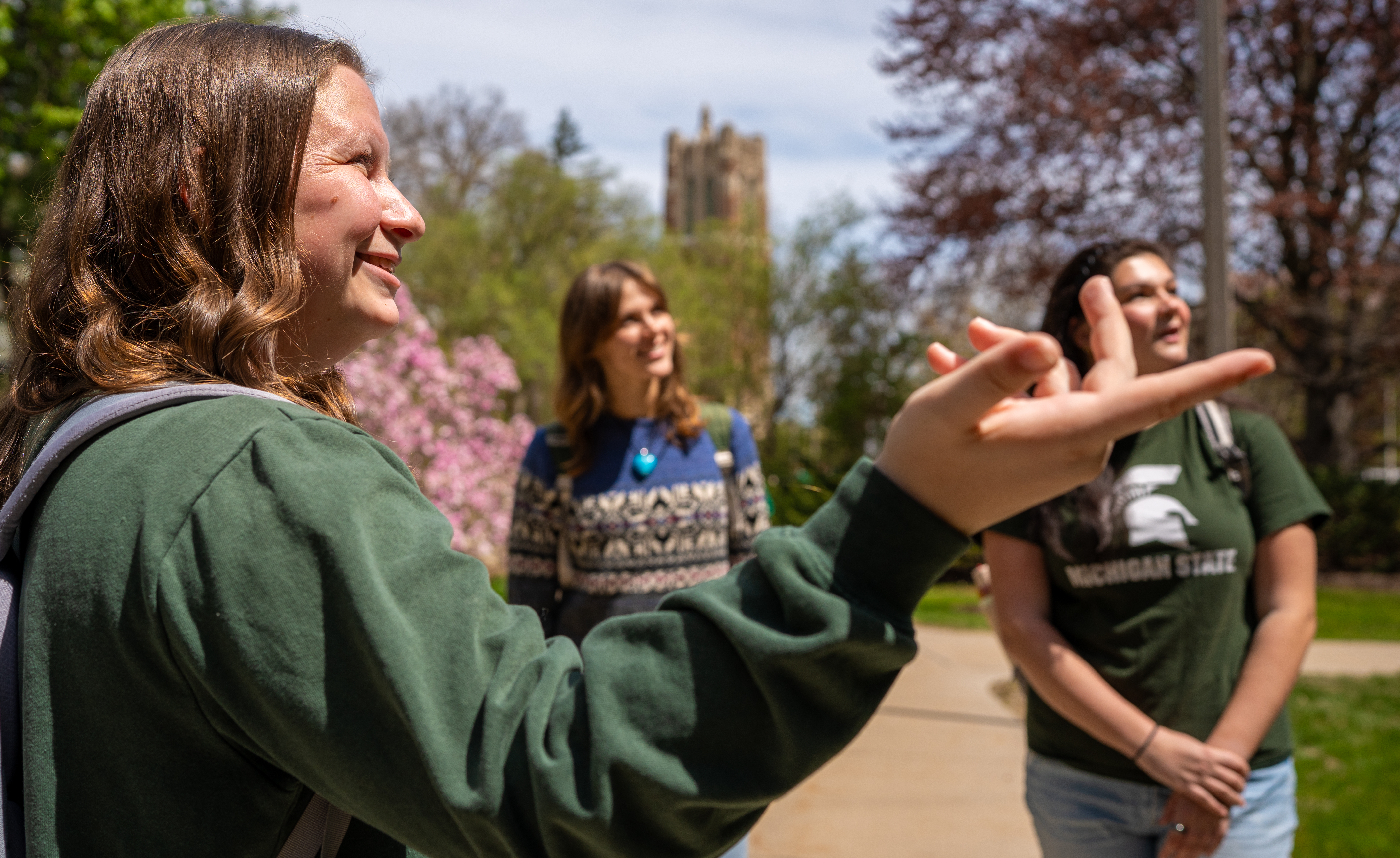 A smiling student tour guide, Breanne Sandstrom, leads a tour on the Michigan State University campus. Two other Spartans listen attentively in the background, with spring trees and Beaumont Tower visible behind them.