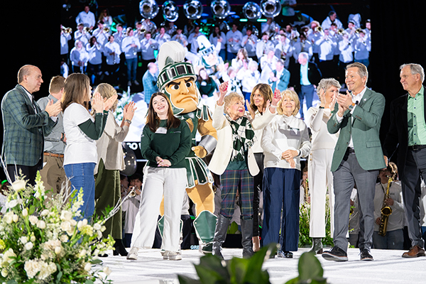 Sparty and a group of Spartans celebrate on stage in front of a large video screen and live band during a campaign event.
