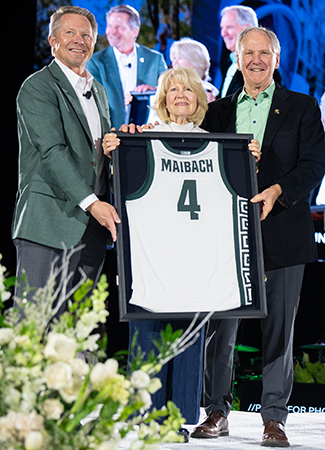 Barb and Ben Maibach stand next to MSU President Guskiewicz as he presents a framed basketball jersey to them