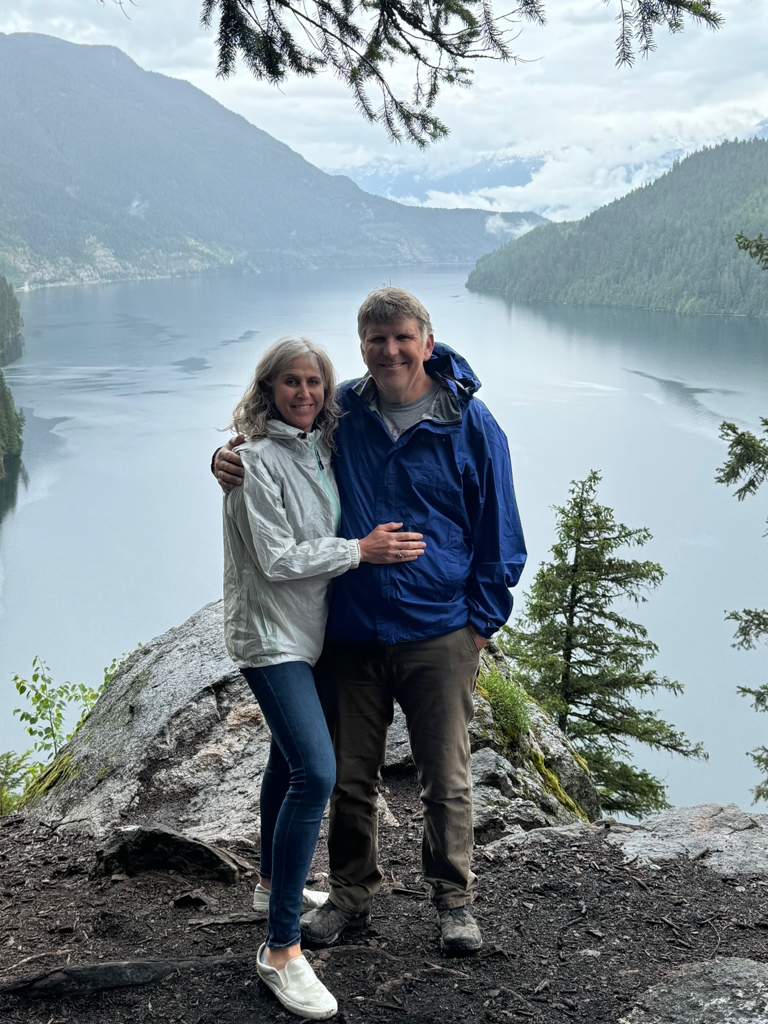 Meryl Katz and Jack Peurach standing together on a scenic overlook, smiling. Behind them is a mountain lake surrounded by evergreen forests and mist-covered peaks.