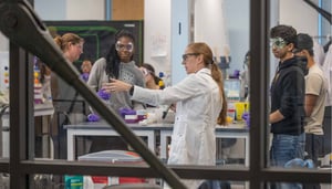 A woman in a lab coat talks to a group of students in a classroom with lab equipment.