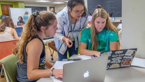 Two students sit at a table with laptops. A third leans over to talk to them.