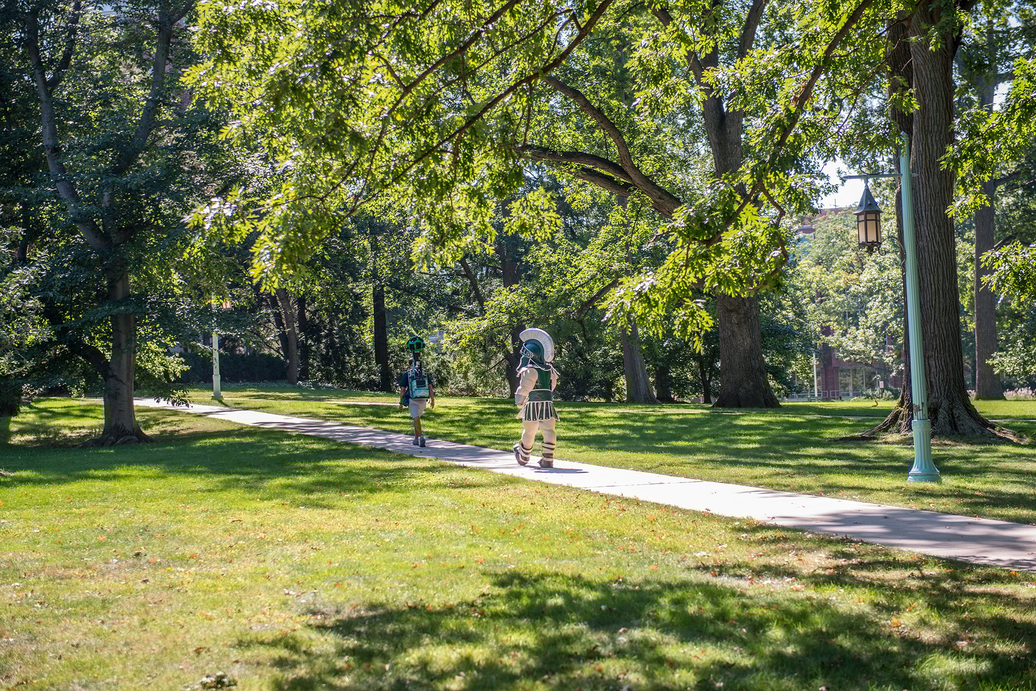  person and Sparty leisurely walk along a park path on campus, immersed in the beauty of the lush landscape.