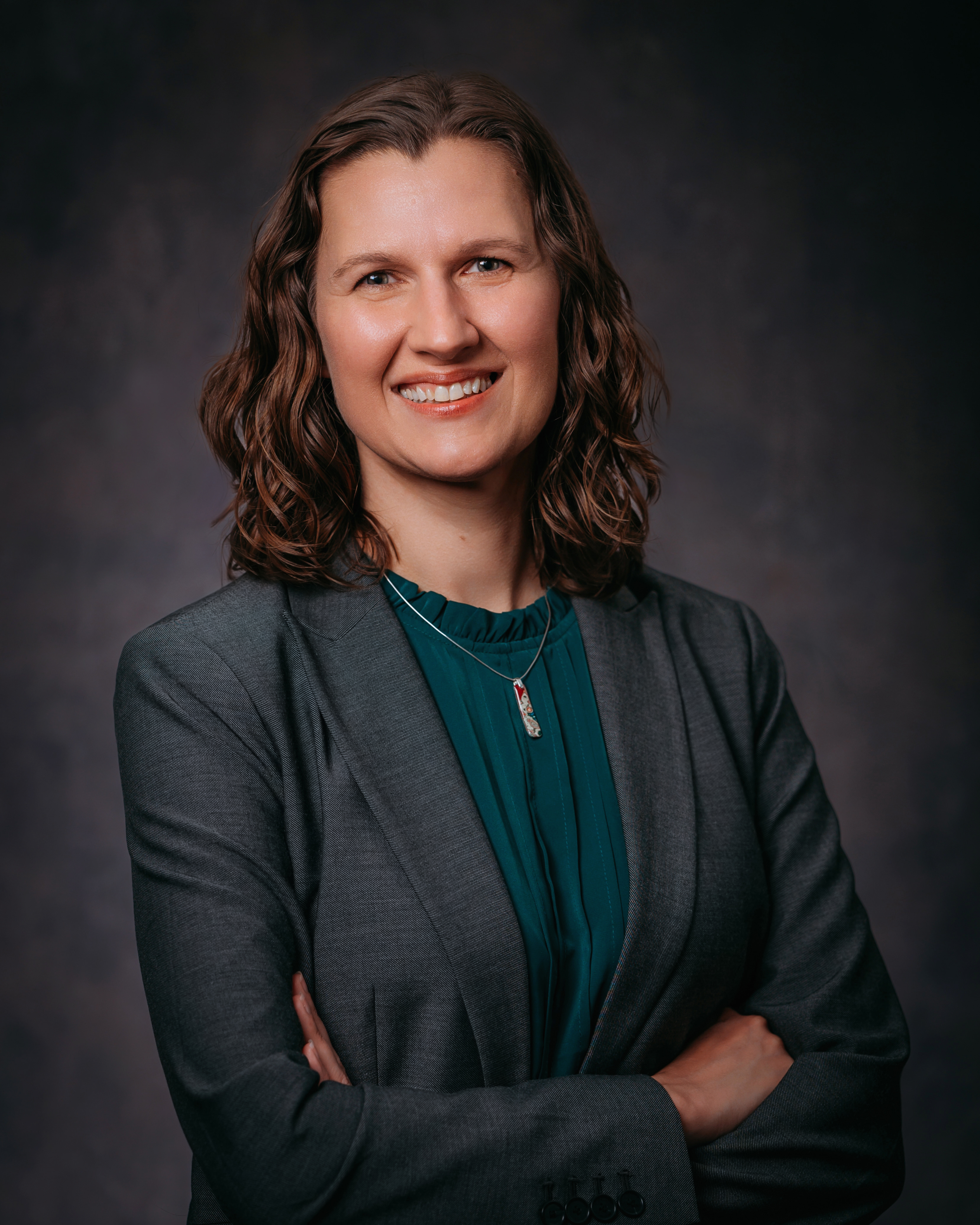 Studio Portrait of Renata Opoczynski wearing a grey blazer over a dark teal top. She has dark curly hair.