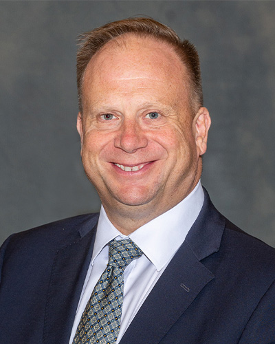 Portrait of Mark Largent. He is wearing a dark colored jacket with a white collared shirt and white and blue tie, his hair is cut short, and he is against a dark grey background smiling at the camera.