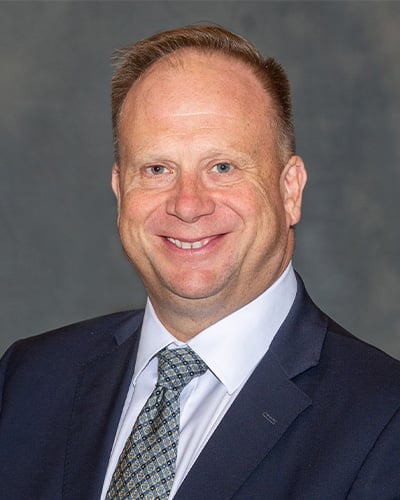 Portrait of Mark Largent. He is wearing a dark colored jacket with a white collared shirt and white and blue tie, his hair is cut short, and he is against a dark grey background smiling at the camera.
