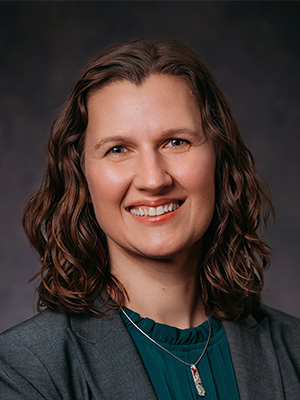 Portrait photo of Renata Opoczynski. She is looking at the camera and smiling, with curly hair wearing a grey blazer and green shirt.