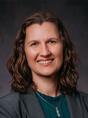 Portrait photo of Renata Opoczynski. She is looking at the camera and smiling, with curly hair wearing a grey blazer and green shirt.