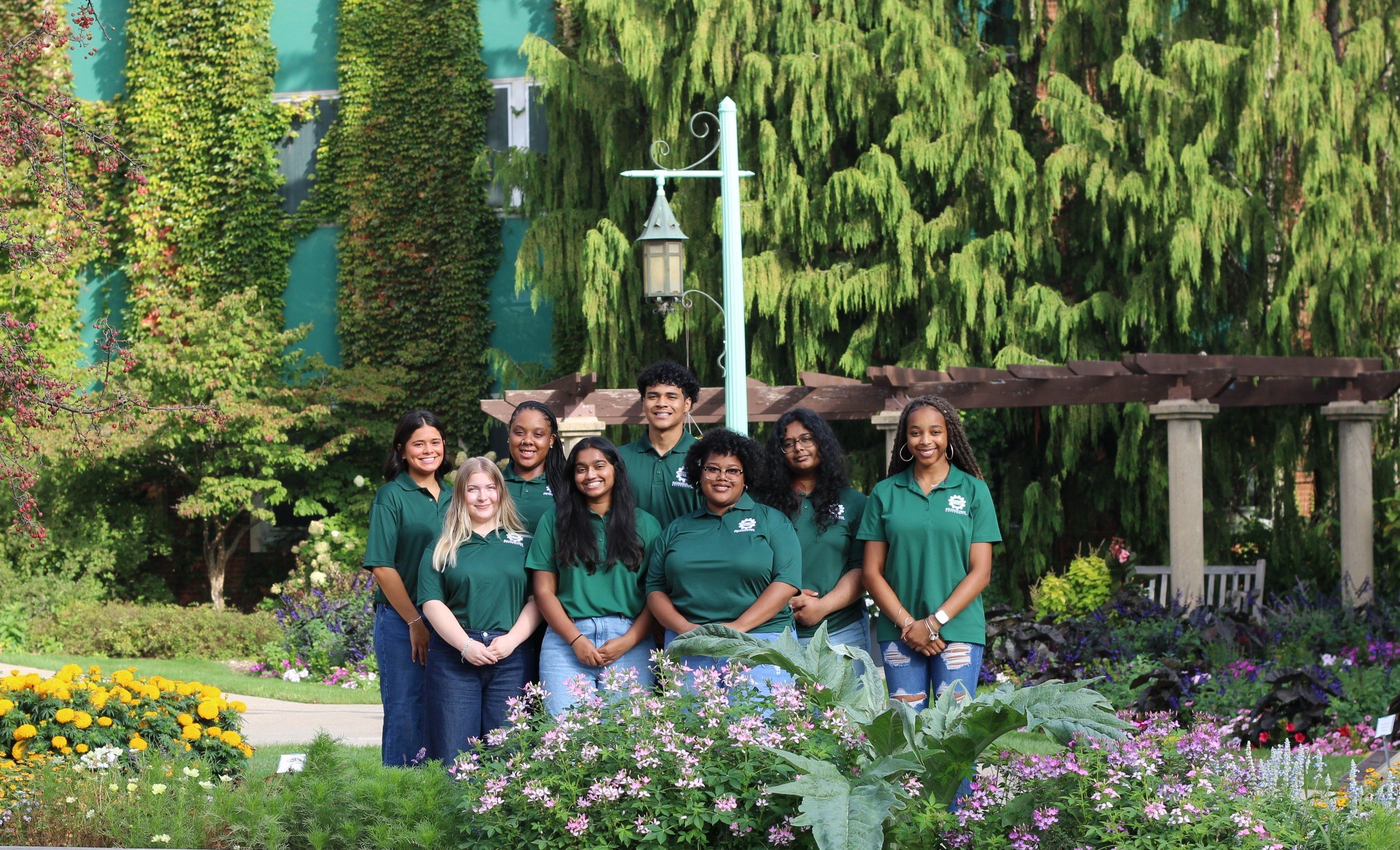 A group of students standing together facing the camera all wearing the same green colored polo shirt, backdropped by trees and a lantern from MSU's campus