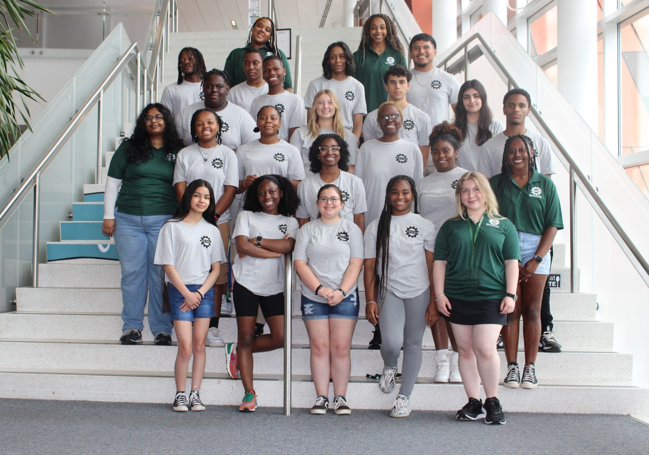 A group of students standing together smiling and facing the camera on a staircase; all people are wearing either the same white or green shirt.