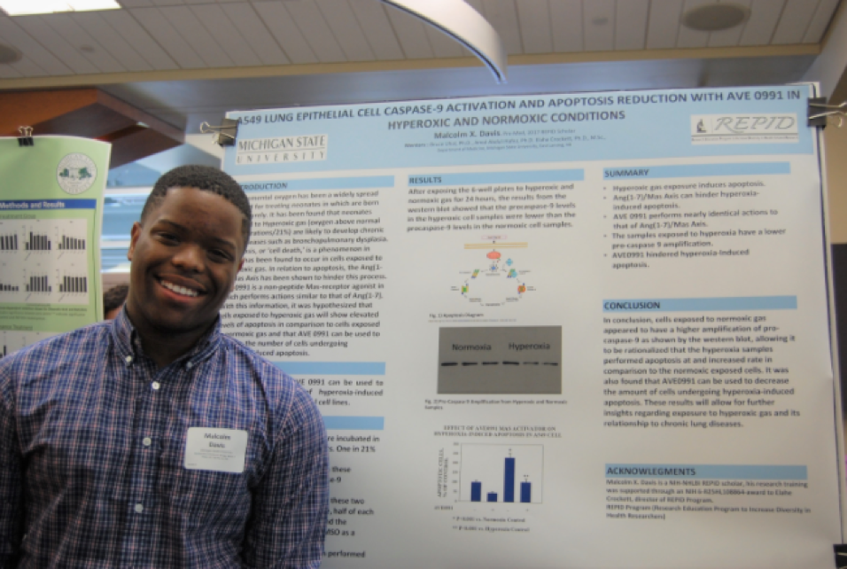 A man stands in front of a research poster smiling widely at the camera.