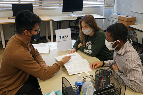 Two students are sitting at one side of the table talking with a person on the other side in front of a TRIO sign.
