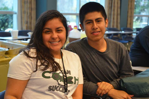 Two students sitting next to one another at the center of the frame smiling while looking at the camera.