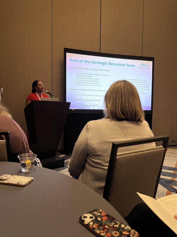 A person stands behind a podium next to a presentation screen and speaks toward the audience