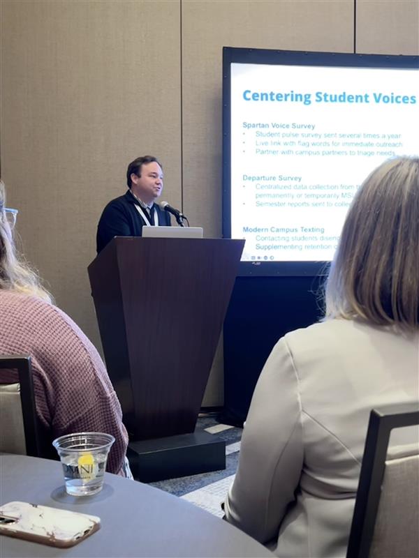 A person stands behind a podium next to a presentation screen and speaks toward the audience