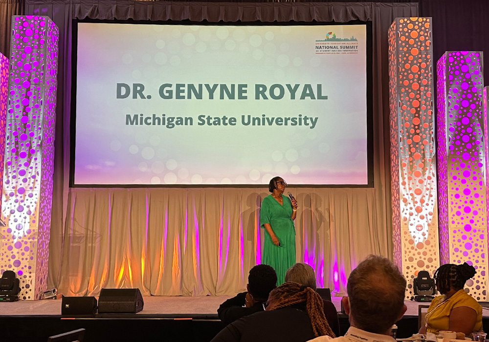 A person in a green dress standing just right of center stage in front of a large screen that reads Dr. Genyne Royal, Michigan State University