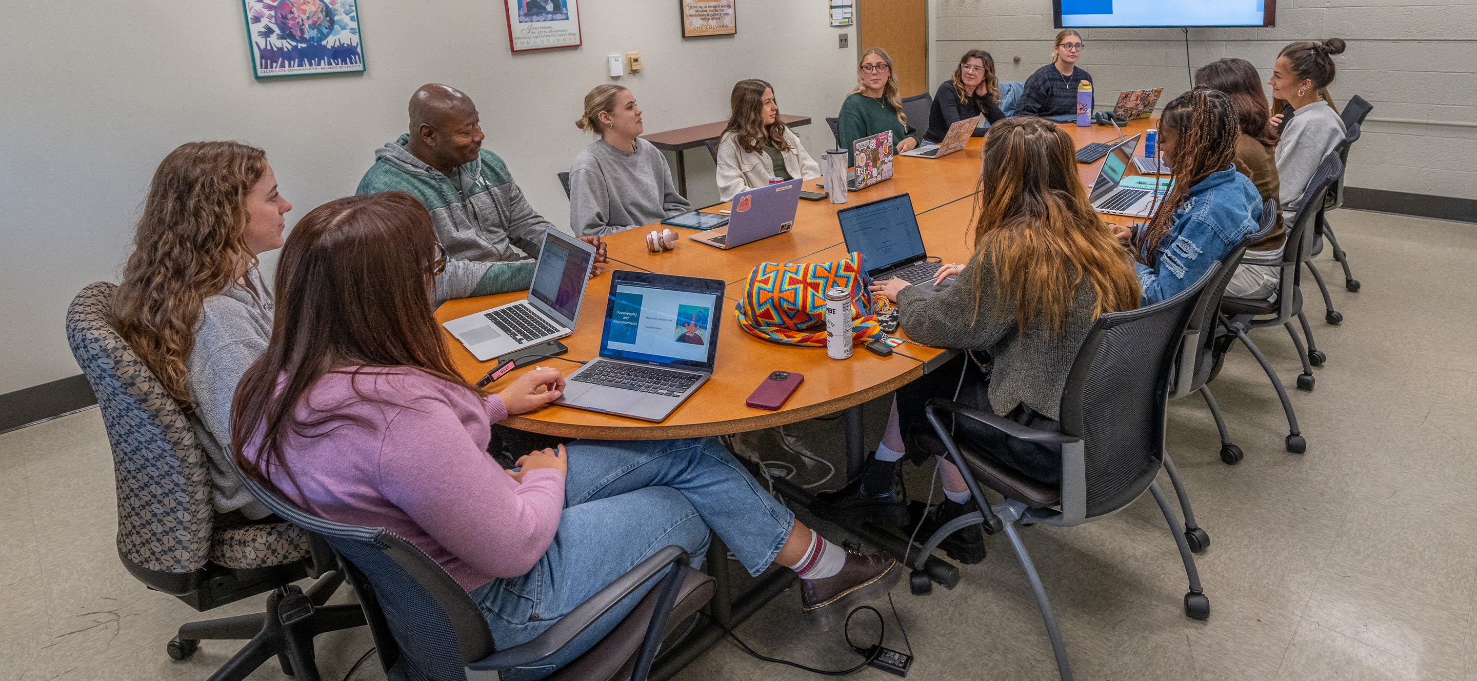 A classroom group sits around a brown oval table with a large screen at one end of the table. Students have books, laptops and other classroom materials spread out on the table.