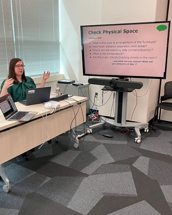 A person sits at a desk with their arms up while speaking next to a large screen displaying an informational slide during the CTLI Setting the Tone from the Start hybrid workshop.