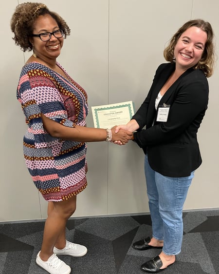 Two people are standing next to each other facing the camera and smiling while shaking hands over a certificate. 