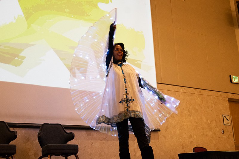 A person stands on a stage in front of a projection screen with one arm lifted up above them as if about to take flight, while wearing a sheer colored, transparent cape.