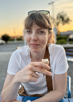 Portrait of person sitting facing and looking into the camera holding an almost finished ice cream cone directly in front of them. They are wearing a white t-shirt and are backdropped by the colors of the setting sun.