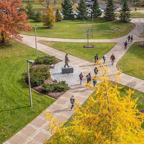 Aerial view of the John Hannah statue in front of MSU's Administration Building. The leaves are changing to fall colors on the trees and students can be seen walking on the sidewalks around the statue. 