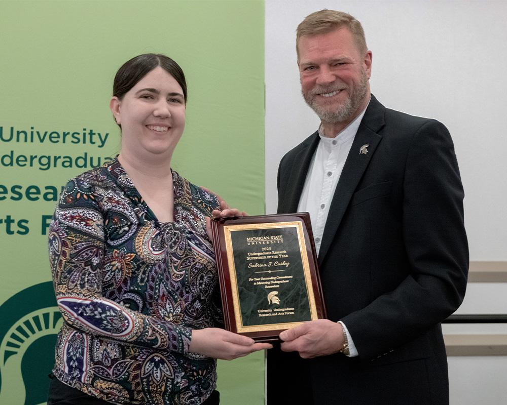 Interim Provost Thomas Jeitschko, wearing a white button-down shirt and a black jacket with a Michigan State University Spartan lapel pin, poses with Sabrina Curley, wearing a shirt with a paisley design, both holding a square plaque honoring Prior as an Undergraduate Research Supervisor of the Year winner.