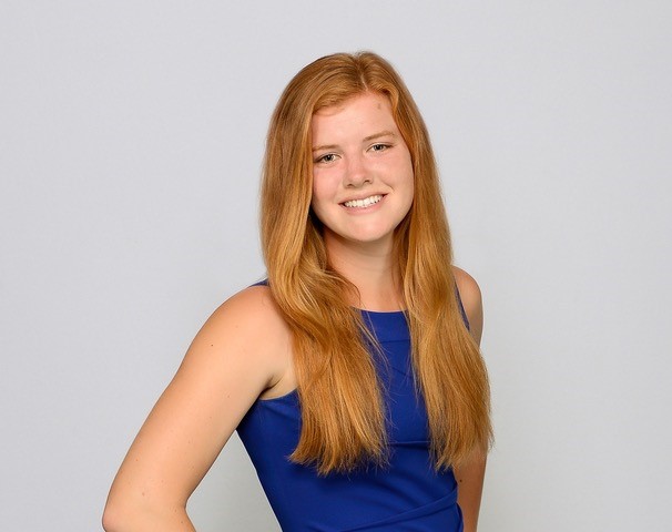 Portrait image from the waist up. She is wearing a blue sleeveless top, with long auburn-red hair down across both shoulders. She is smiling and half turned looking at the camera standing against a white backdrop. 