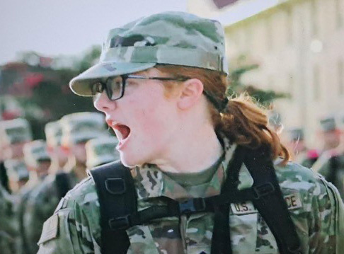 Image of a cadet from the chest up wearing glasses, a hat and a backpack, dressed in fatigues. Her head is turned to the side and with her mouth open speaking to the cadets in formation behind her.