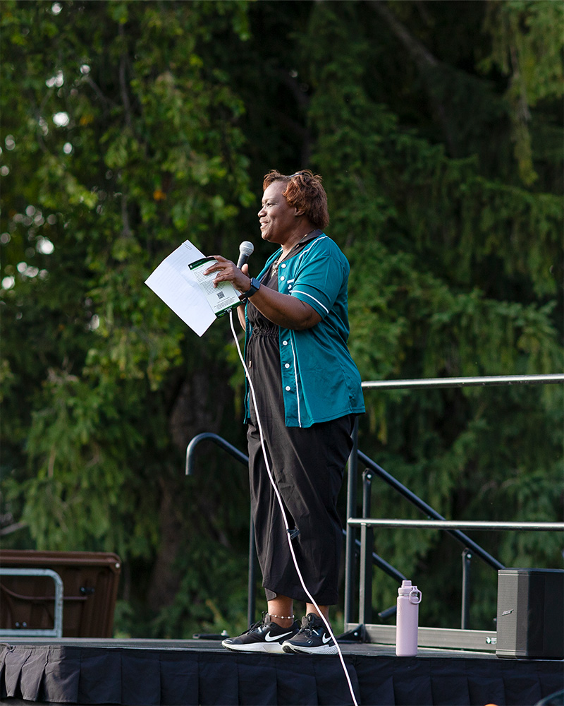 A person wearing a blue shirt and black pants stands on a stage speaking into a microphone while holding a paper in their hand, backdropped by green foliage and trees. 