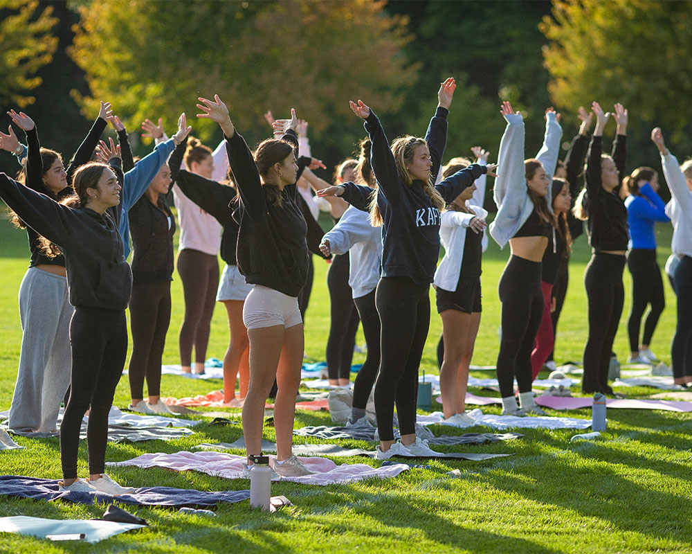 A group of people standing in rows on yoga mats stand with their arms stretched out above them toward the sky. They are standing in a green, grassy field on a sunny day. 