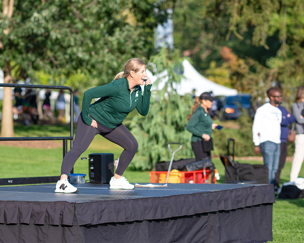 A speaker wearing black yoga pants and green, long sleeved shirt, stands on a black stage looking out in front while leaning their body weight onto their left leg.
