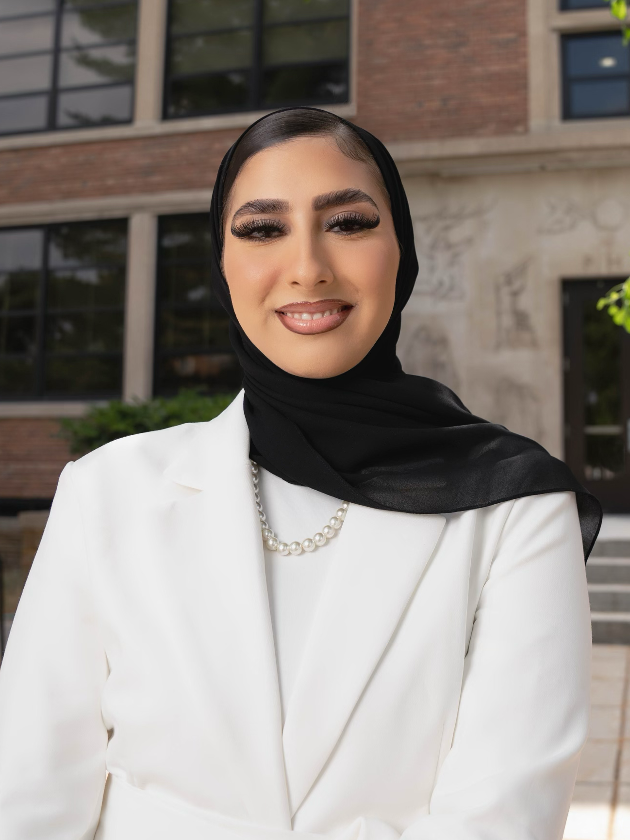 Mariam Turkey wearing a white jacket and black head covering smiles while standing outside a campus building
