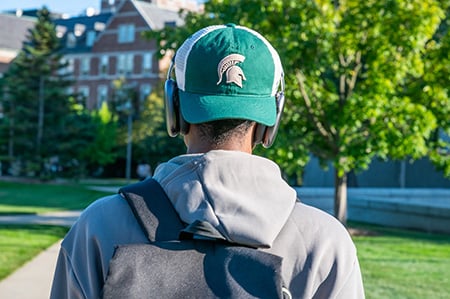 The back of a student wearing a Spartan helmet hat with the student framed by green trees and the sidewalk ahead.