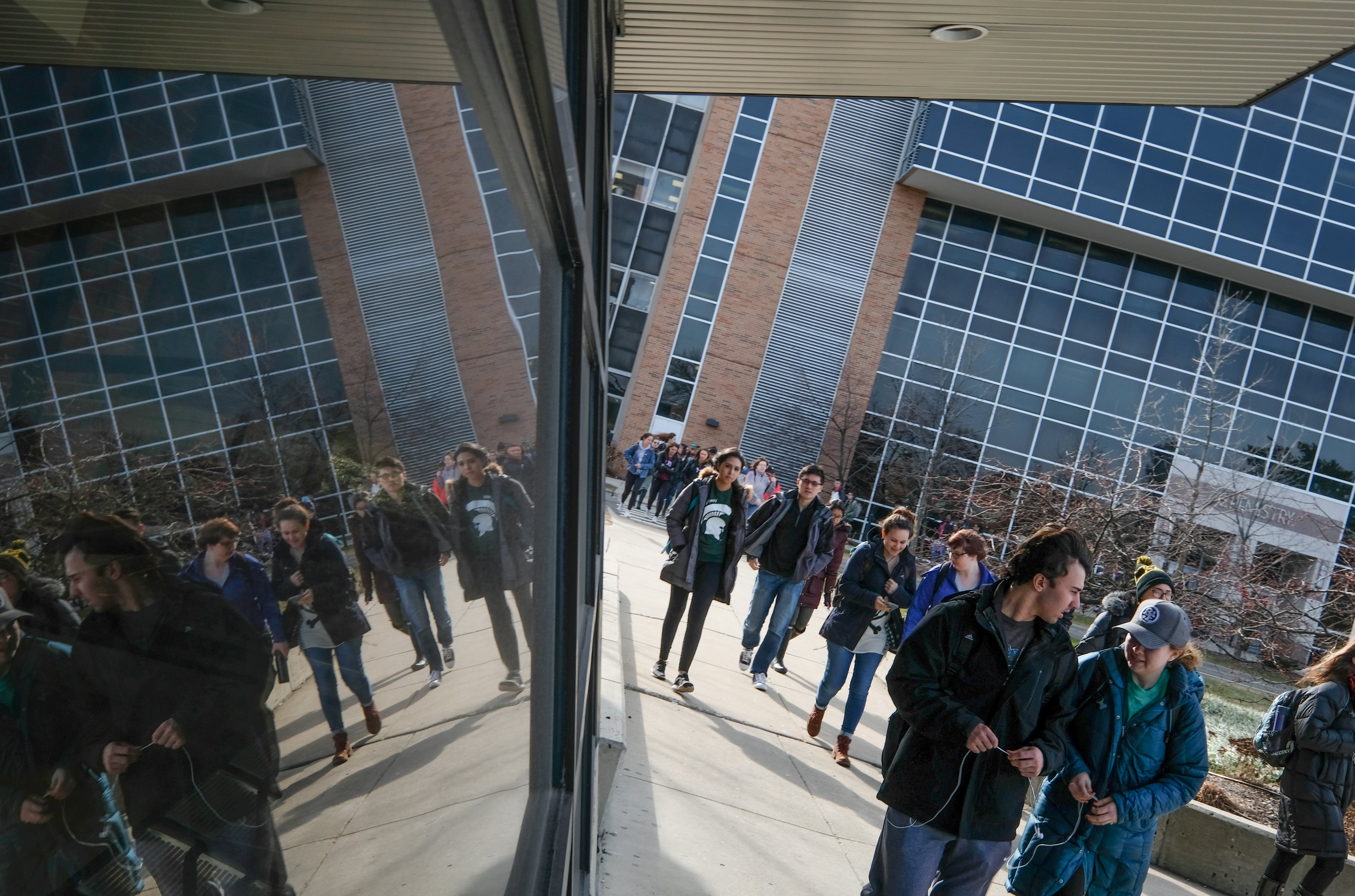 Students walk past the main bus station in central campus, their reflections visible in the windows of the covered waiting area.