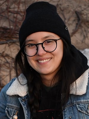 Portrait image. The individual is looking at the camera smiling, with long dark hard, glasses, while wearing a dark, knitted hat and coat with a plush collar.