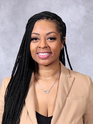 Portrait image. The individual is wearing a tan suit jacket and black shirt, with long hair draped over their right shoulder against a grey background.